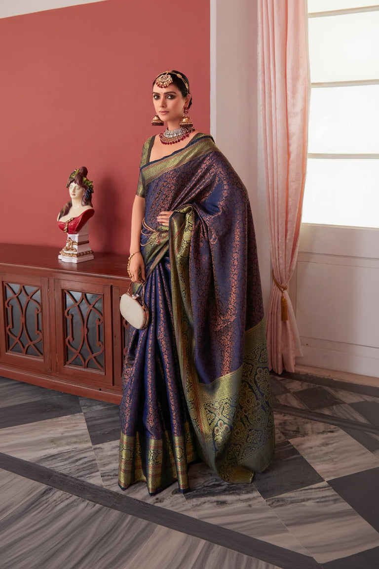 Woman in a traditional saree standing in a room with a decorative cabinet and pink curtains.