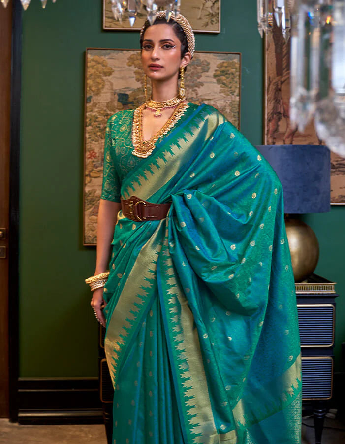 Woman in a teal saree with gold patterns standing in an elegant room.