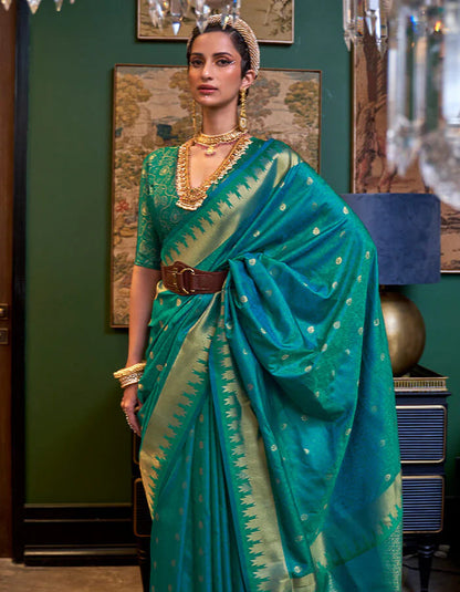 Woman in a teal saree with gold patterns standing in an elegant room.