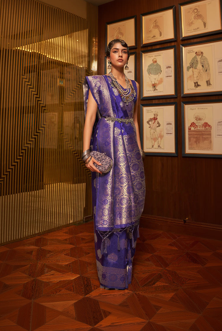 Woman in a traditional purple outfit with jewelry, holding a clutch, in an indoor setting.