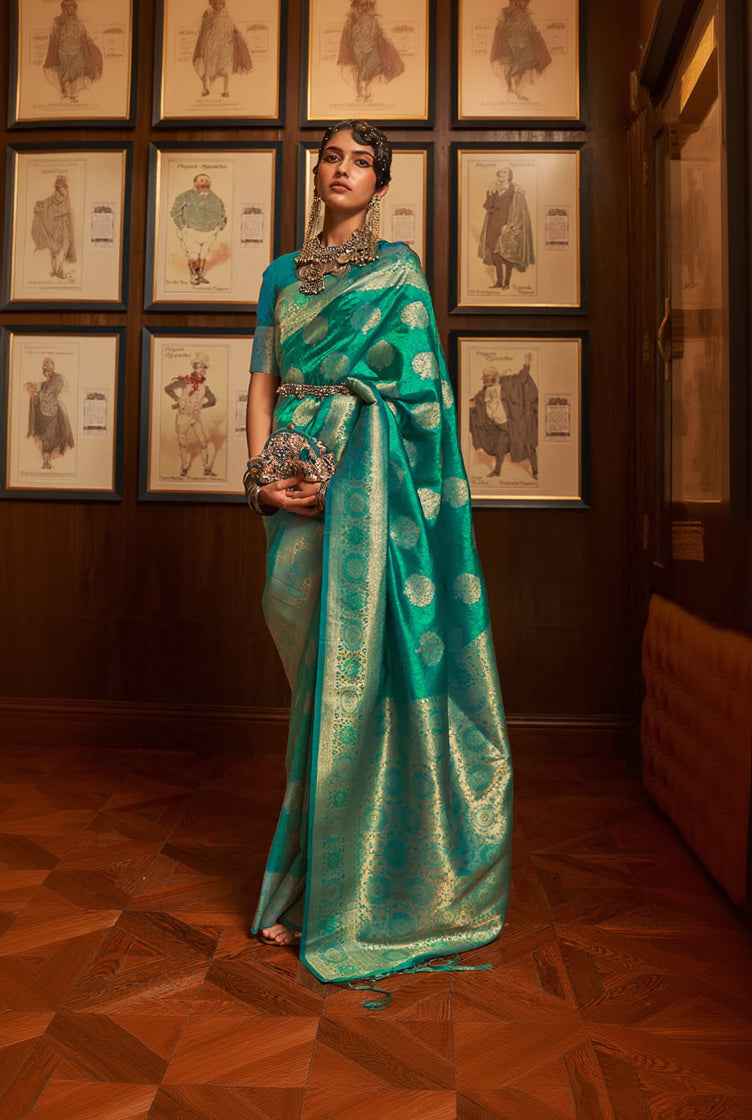 Woman in a green saree with gold patterns standing in front of framed pictures on a wall.